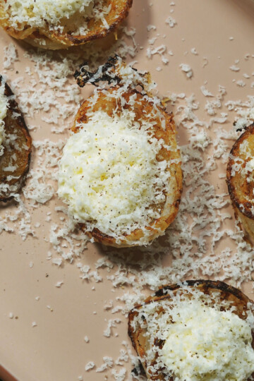Five pieces of toasted bread topped with creamy white cheese and freshly grated cheese are arranged on a beige plate. The cheese is sprinkled generously, leaving shavings scattered on the plate.