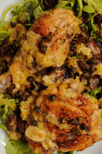 A close-up of roasted chicken pieces topped with a creamy, caper-studded sauce, served on a bed of fresh arugula and caramelized onions.