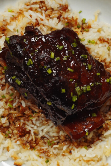 A close-up of glazed short ribs garnished with chopped chives, served on a bed of white rice drizzled with sauce.