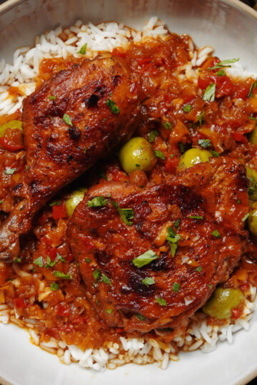 A plate of white rice topped with two pieces of braised chicken in a rich tomato-based sauce, garnished with chopped herbs and green olives.