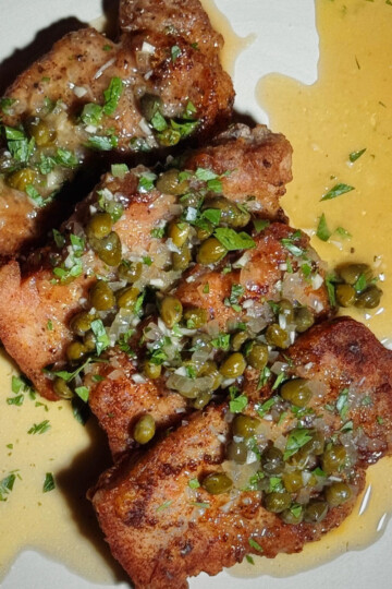 Three pieces of breaded fish topped with capers, chopped herbs, and a lemon butter sauce, served on a white plate with sauce drizzled around them.