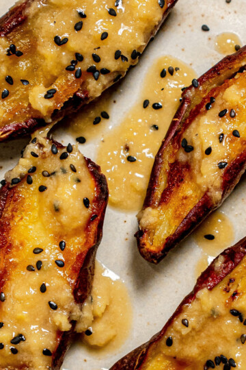 Four roasted sweet potato halves topped with a creamy sauce and sprinkled with black sesame seeds, arranged on a light-colored plate.