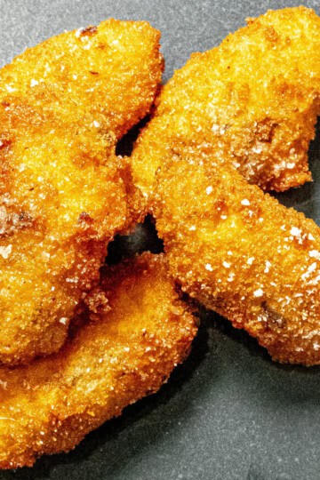 Four crispy, golden-brown breaded chicken tenders with visible salt flakes on a dark gray surface.