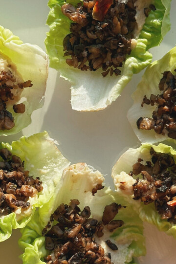 Six lettuce leaves arranged on a white surface, each filled with a savory mixture of finely chopped mushrooms and vegetables, topped with small pieces of red chili. Sunlight casts gentle shadows across the dish.