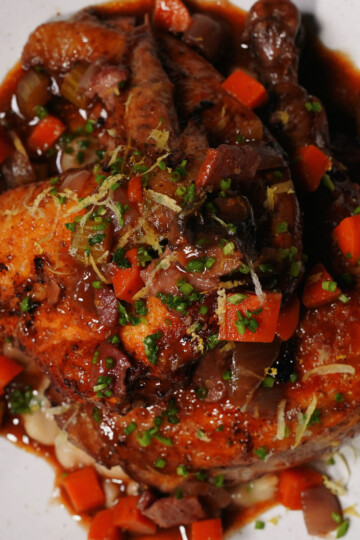 A roasted chicken served on a white plate, topped with a rich brown sauce, diced carrots, celery, mushrooms, herbs, and lemon zest, with visible pieces of vegetables surrounding the chicken.