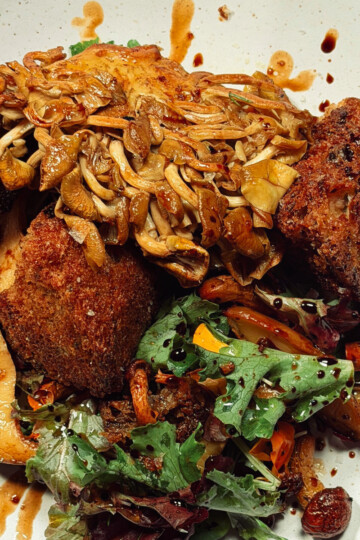 A plate of crispy fried tofu topped with sautéed sliced mushrooms, served with a fresh mixed greens salad, cherry tomatoes, roasted carrots, and a balsamic glaze drizzle.