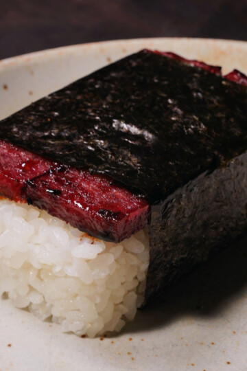 A piece of white rice topped with a slice of cooked Spam, wrapped together with a strip of dark seaweed, served on a light-colored ceramic plate.