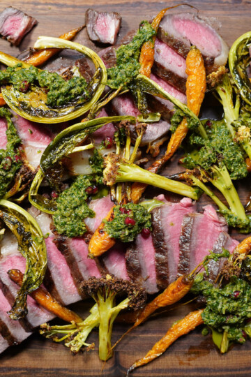 Sliced medium-rare steak topped with green chimichurri sauce, surrounded by roasted baby carrots, broccoli, and charred scallions, all arranged on a wooden board.