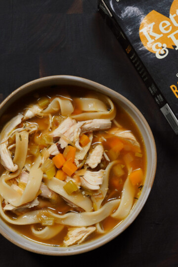 A bowl of chicken noodle soup with wide noodles, shredded chicken, carrots, and celery sits on a dark surface next to a carton of Kettle & Fire Classic Chicken Bone Broth.