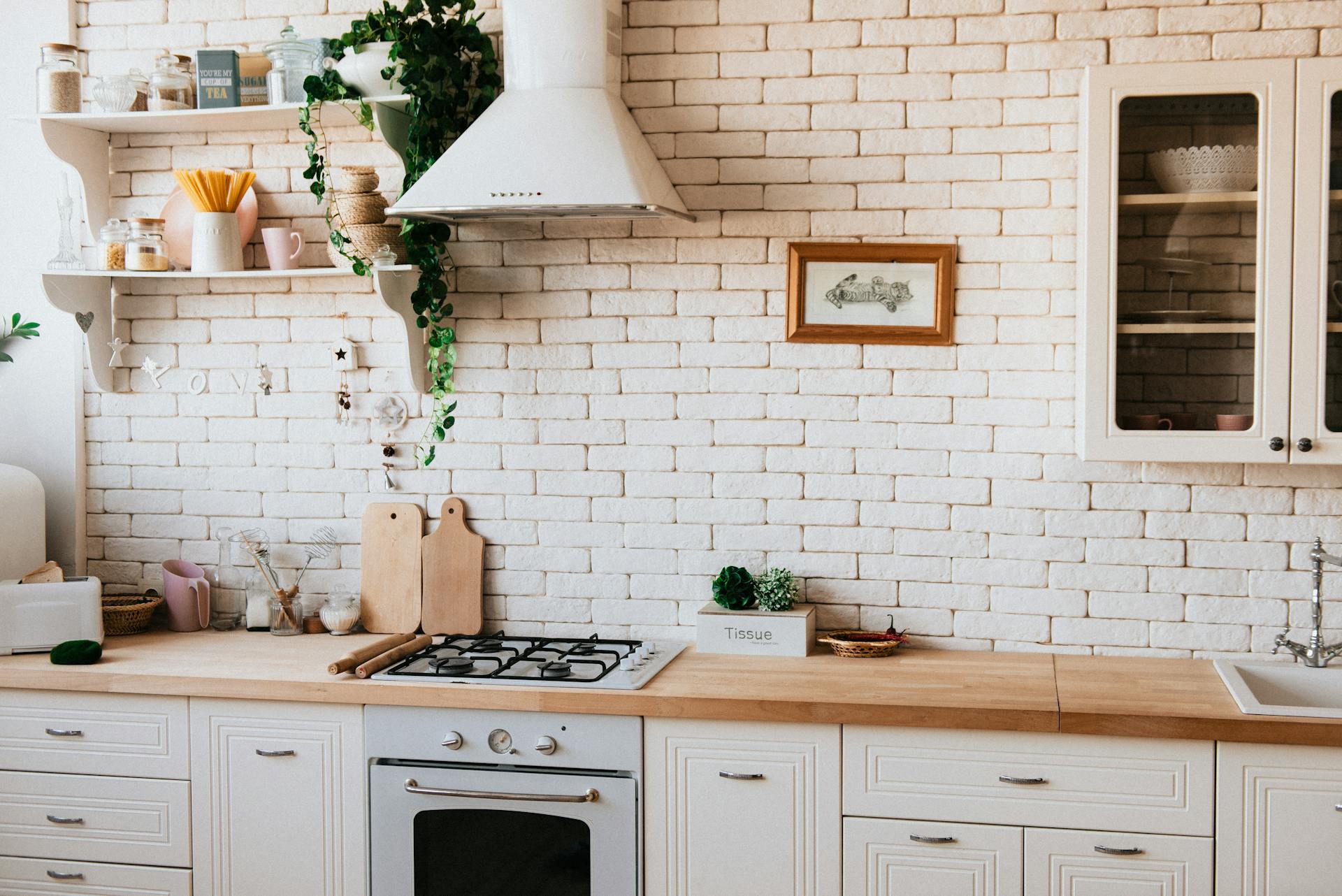 A bright, cozy kitchen with white cabinets, wooden countertops, a gas stove, open shelves with dishes and plants, and a white brick wall decorated with a framed picture and greenery.