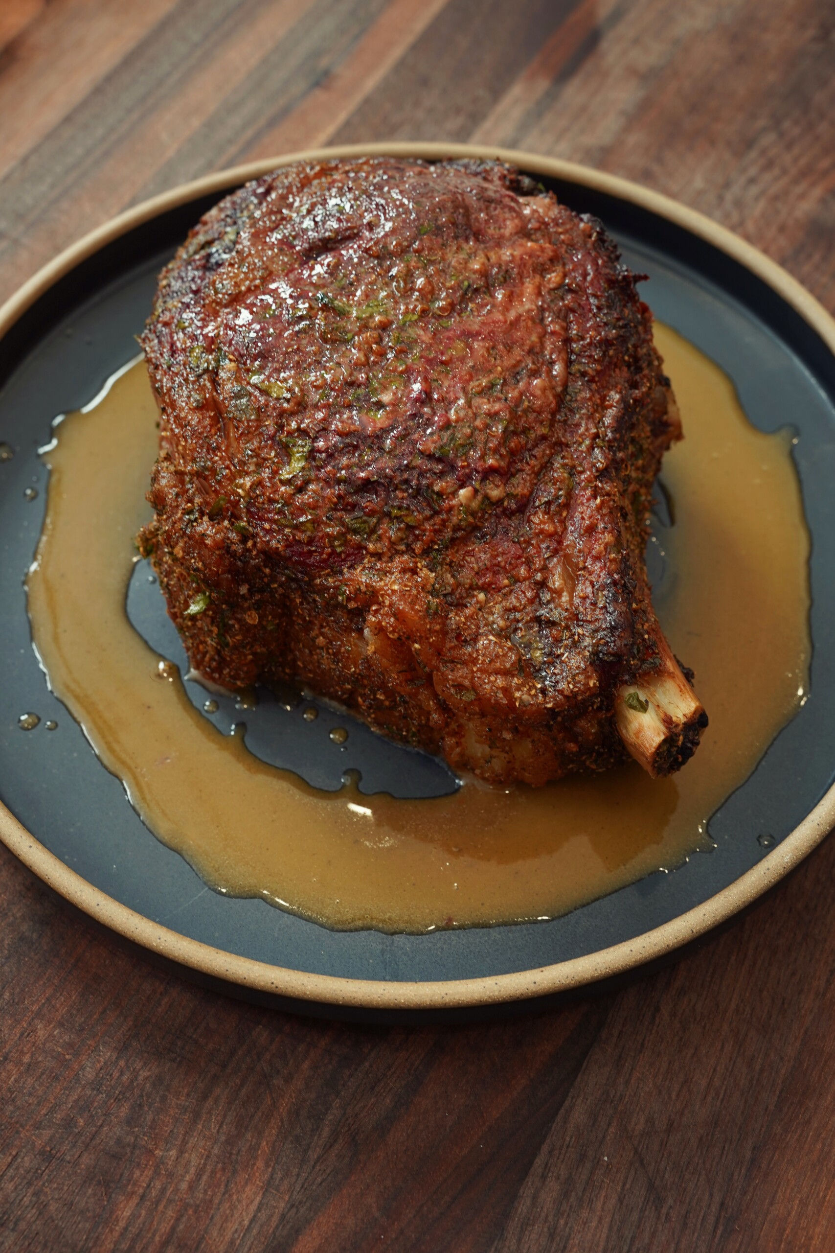A cooked bone-in rib roast sits on a round black plate, surrounded by meat juices, on a wooden surface. The roast is seasoned and has a crispy, browned exterior.