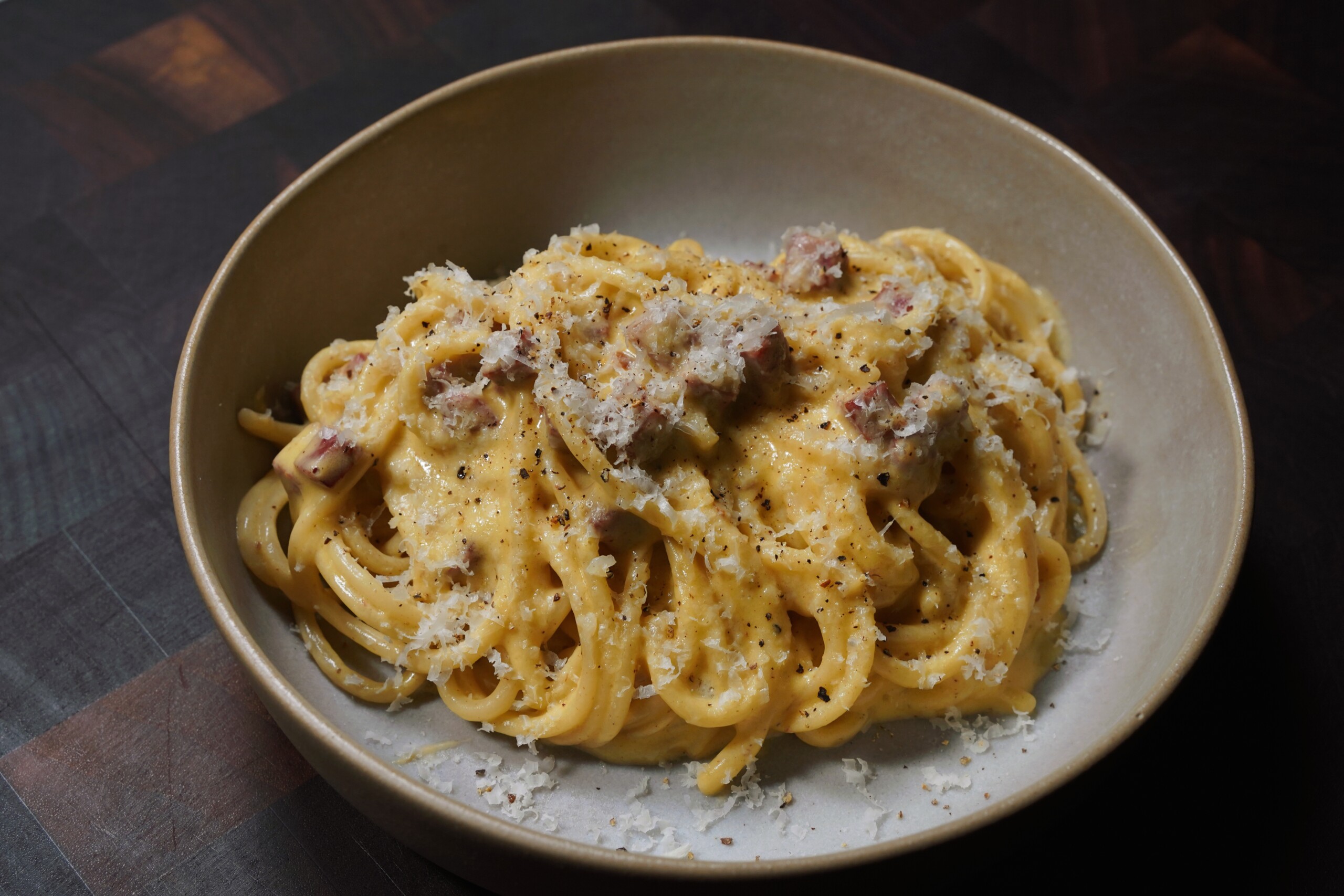 A bowl of spaghetti carbonara topped with grated cheese and cracked black pepper, featuring creamy sauce and pieces of pancetta on a dark wooden surface.