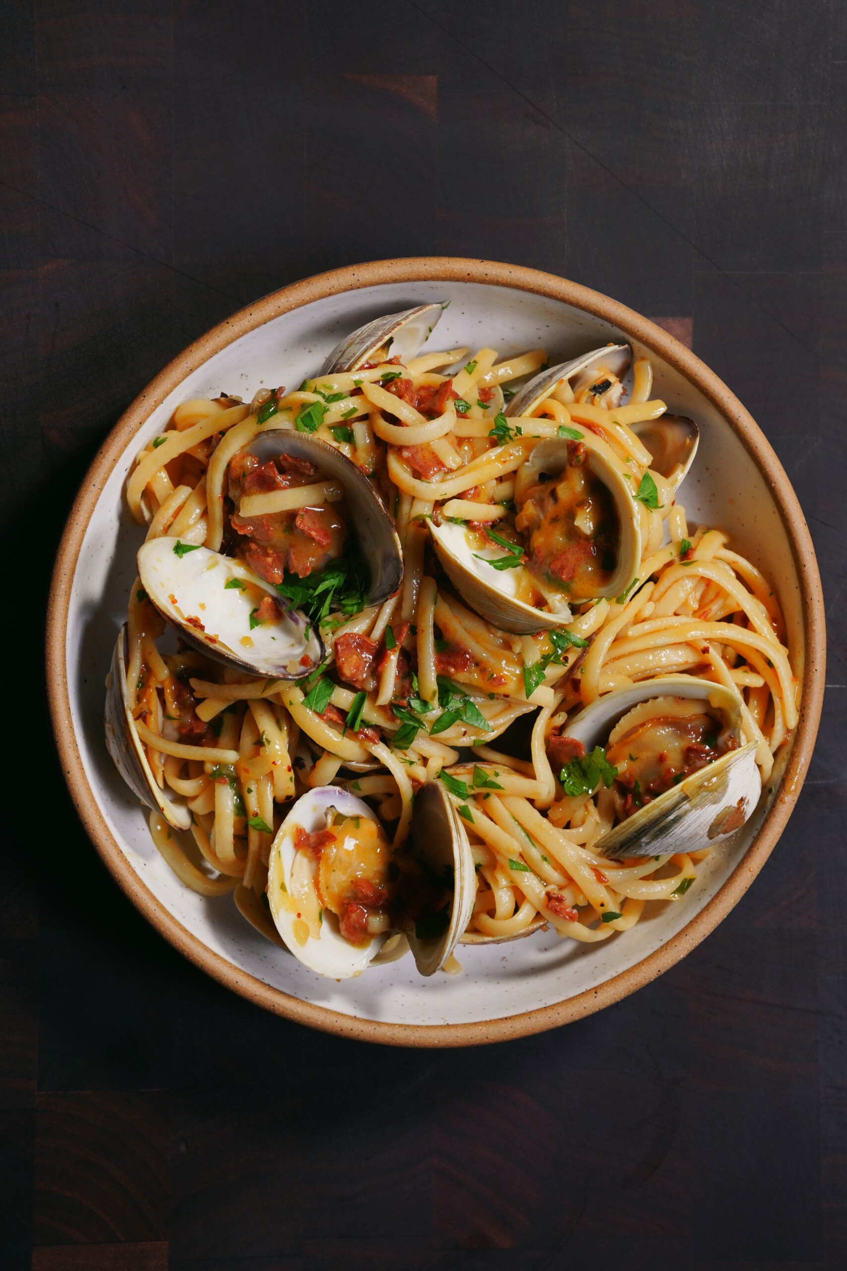A plate of spaghetti with clams in shells, topped with chopped herbs and a light tomato-based sauce, served on a dark wooden surface.