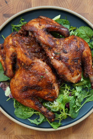 A whole roasted spatchcock chicken with crispy, seasoned skin is served on a bed of fresh mixed greens on a round black plate, placed on a wooden surface.