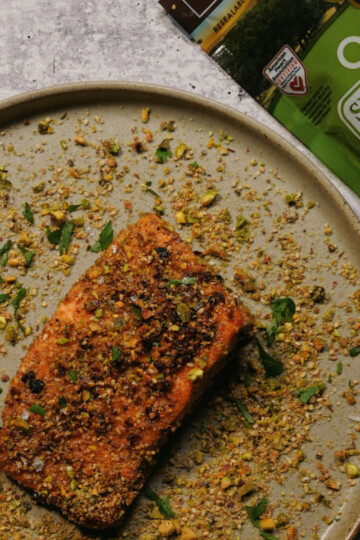 A piece of cooked salmon topped with crushed pistachios on a beige plate, with chopped herbs and pistachio crumbs scattered around. A bag of organic shelled pistachios is partly visible in the corner.