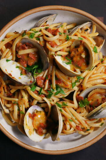 A bowl of linguine pasta with clams in their shells, garnished with chopped parsley and pieces of bacon or pancetta, served in a white dish with a brown rim on a dark wooden surface.