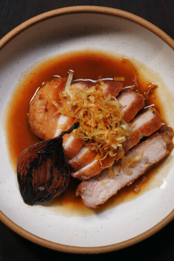 A round plate with sliced roasted meat in brown sauce, topped with crispy fried strips and served with a charred onion and a small round potato on the side.