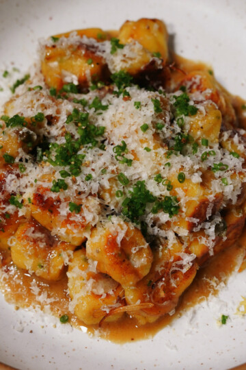 A plate of gnocchi topped with grated cheese, chopped chives, and brown sauce, served on a white ceramic dish with a speckled rim.