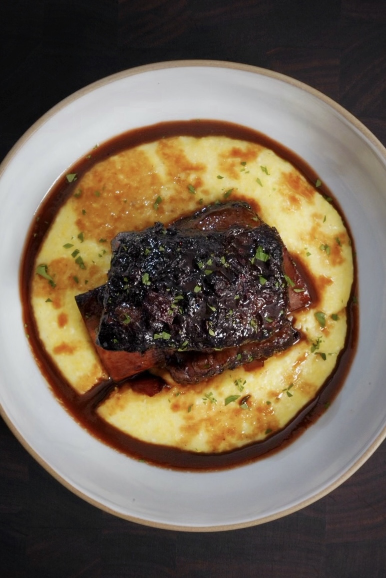 A white bowl of creamy yellow polenta topped with a piece of glazed braised meat, garnished with chopped herbs and surrounded by a rich brown sauce.