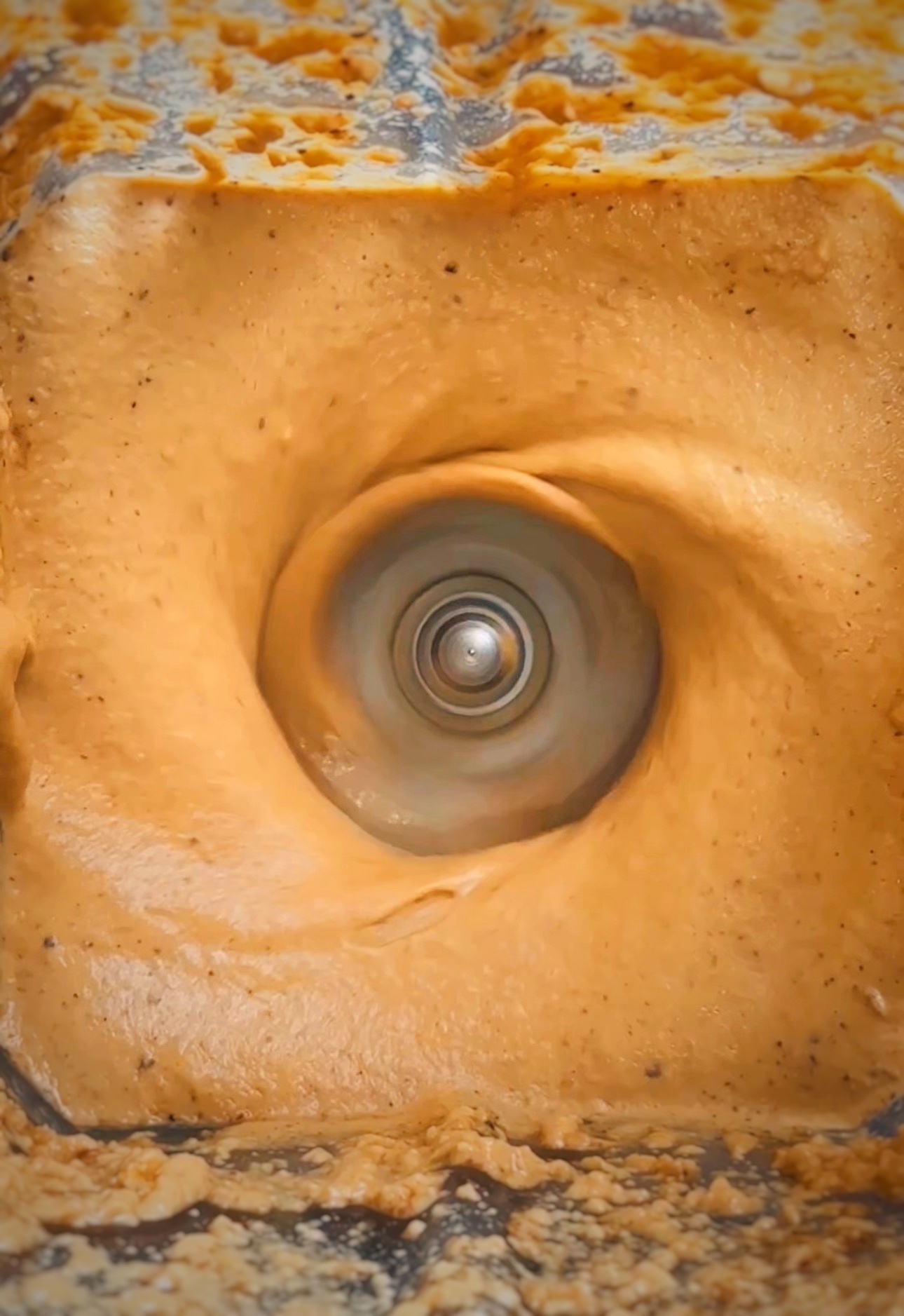 Close-up view of a blender from above, showing a creamy light brown mixture swirling around the spinning blade in the center. The mixture appears smooth and thick, forming a vortex as it blends.