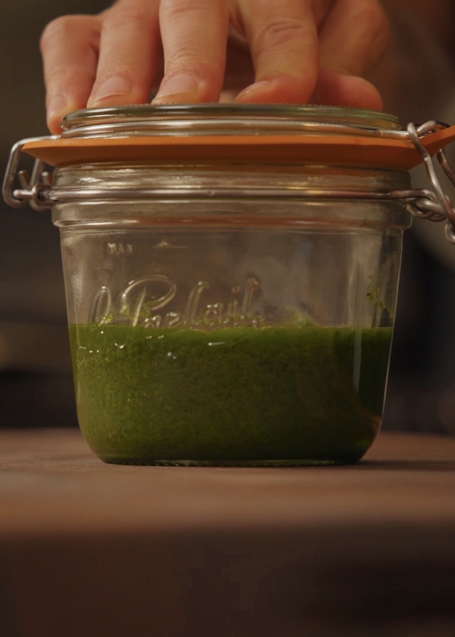 A hand closing the lid of a glass jar containing a small amount of bright green sauce or paste on a wooden surface.