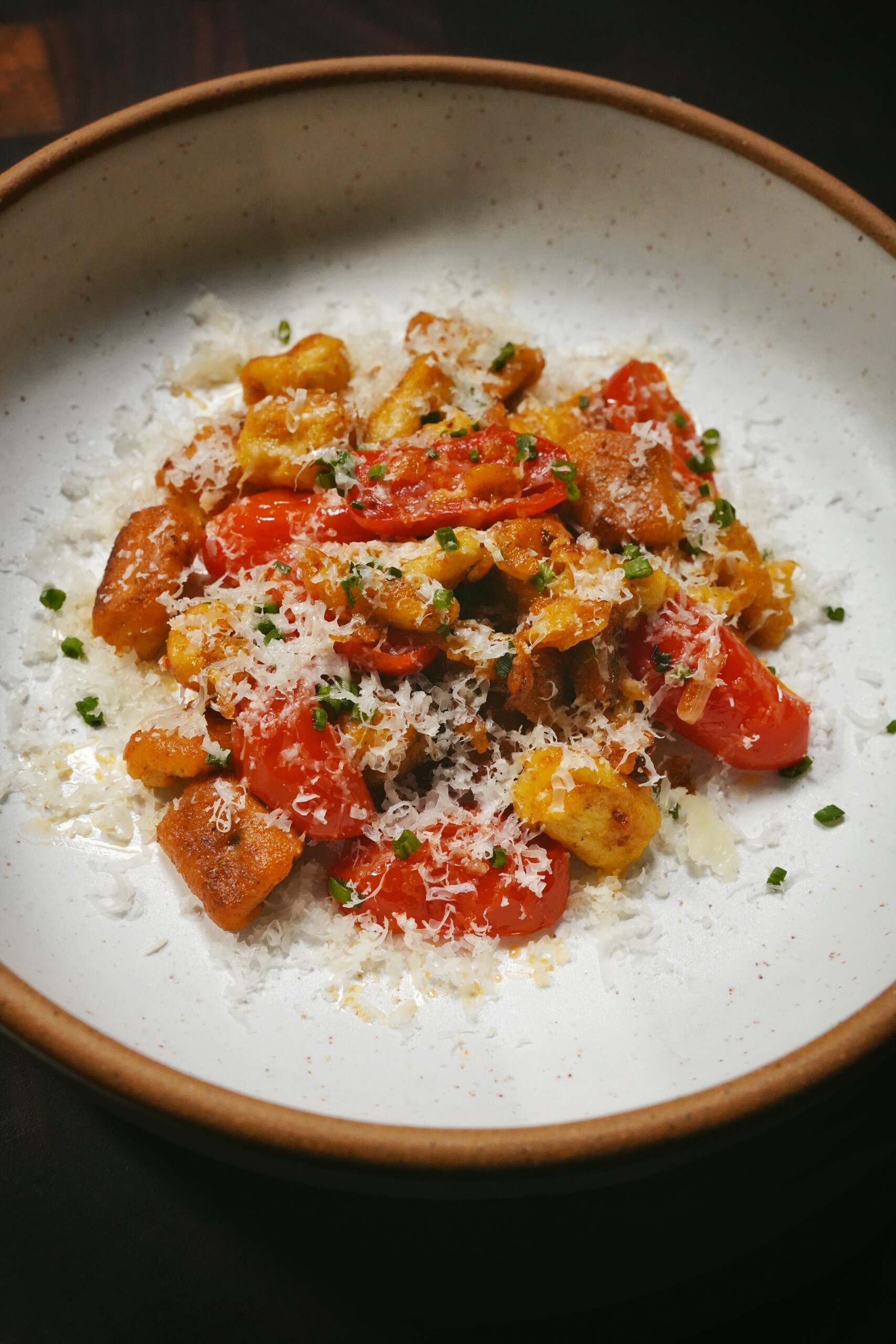 A ceramic bowl filled with gnocchi, cherry tomatoes, chopped herbs, and topped with grated cheese, presented on a dark surface.