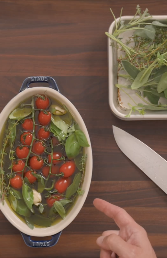 A hand points toward an oval dish filled with vine tomatoes, garlic, basil, and olive oil. Beside it, a tray holds fresh herbs. A large kitchen knife rests on the wooden counter.