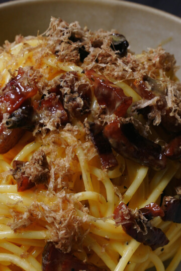 A close-up of a bowl of spaghetti topped with crispy bacon pieces and finely grated truffle, served on a beige plate.