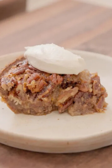 A slice of pecan pie topped with a dollop of whipped cream sits on a simple, round, beige plate on a wooden surface.