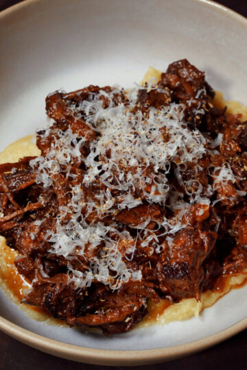 A bowl of creamy polenta topped with shredded braised meat in a rich sauce, finished with a generous amount of finely grated cheese.