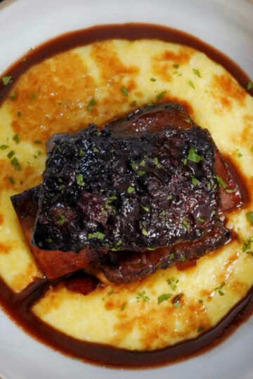 A bowl of creamy yellow polenta topped with braised short ribs in a dark, glossy sauce, garnished with chopped herbs.