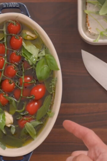 An oval baking dish with cherry tomatoes on the vine, garlic, fresh herbs, and olive oil, sits on a wooden surface. A large knife and a small tray with more herbs are nearby, as a hand points toward the dish.
