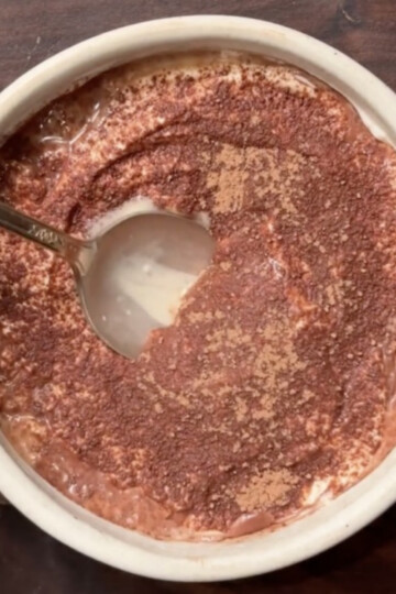 A bowl of chocolate oatmeal topped with cocoa powder sits on a dark wooden surface. A spoon has scooped out a portion, revealing the creamy texture underneath.