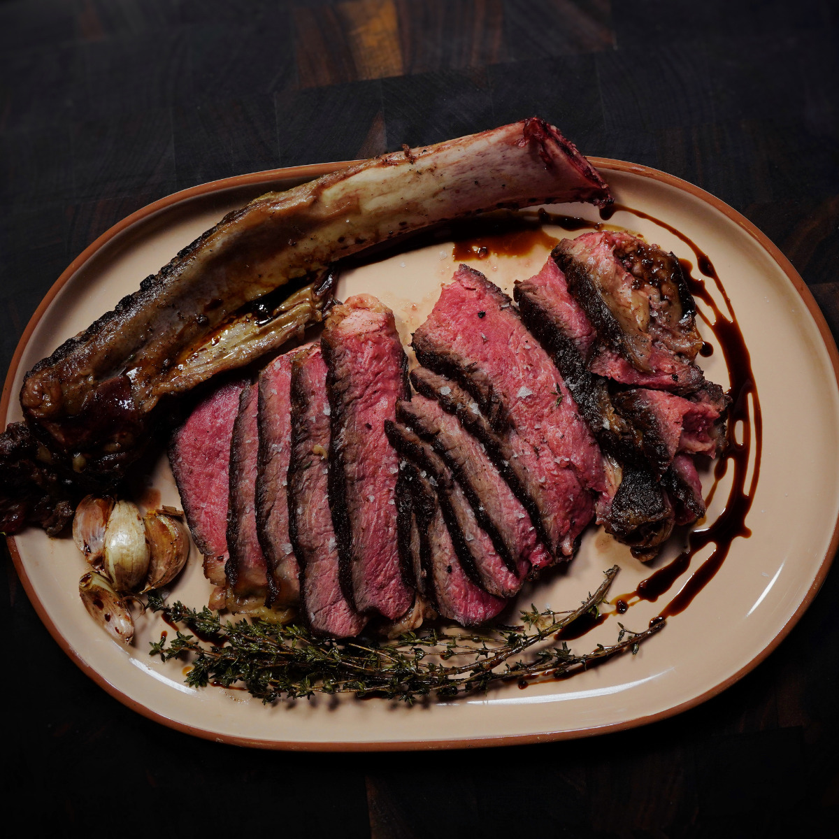 A sliced, medium-rare tomahawk steak is served on a beige plate with roasted garlic, sprigs of thyme, and a drizzle of sauce, set against a dark wooden background.