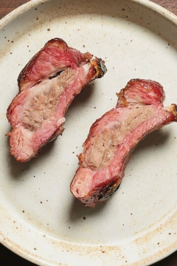 Two pieces of cooked, bone-in meat, likely ribs, with a pinkish interior and browned edges, are placed on a round, off-white ceramic plate with a subtle brown rim, set on a wooden surface.