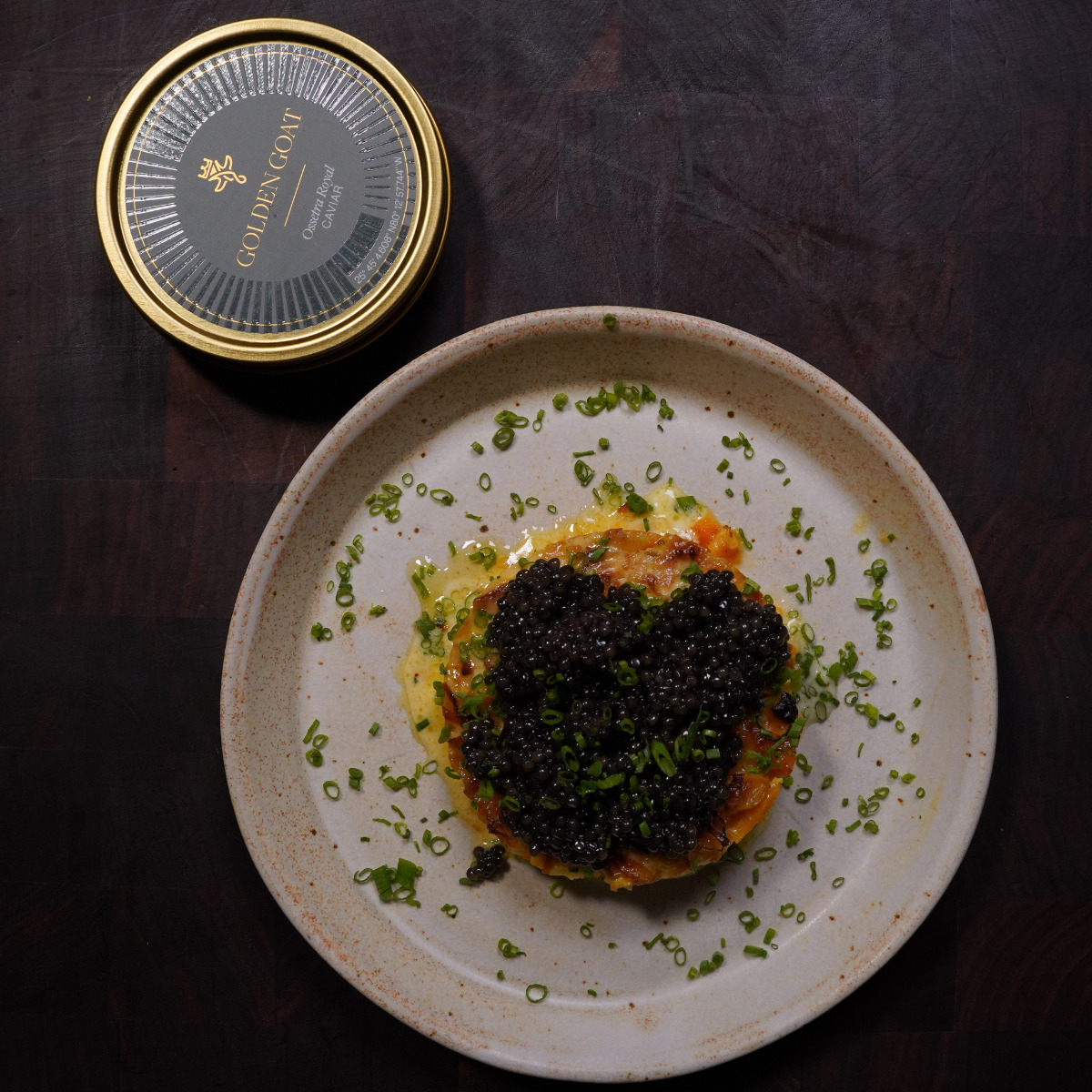 A ceramic plate with a gourmet dish topped with black caviar and chopped chives, next to an open tin labeled Golden Icon Ossetra Caviar on a dark wooden surface.