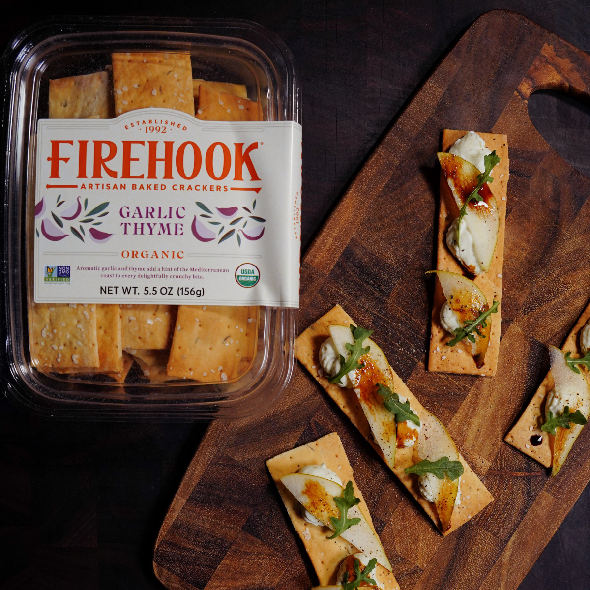 A package of Firehook Garlic Thyme organic artisan baked crackers is on a wooden surface next to several crackers topped with cheese, herbs, and seasoning.