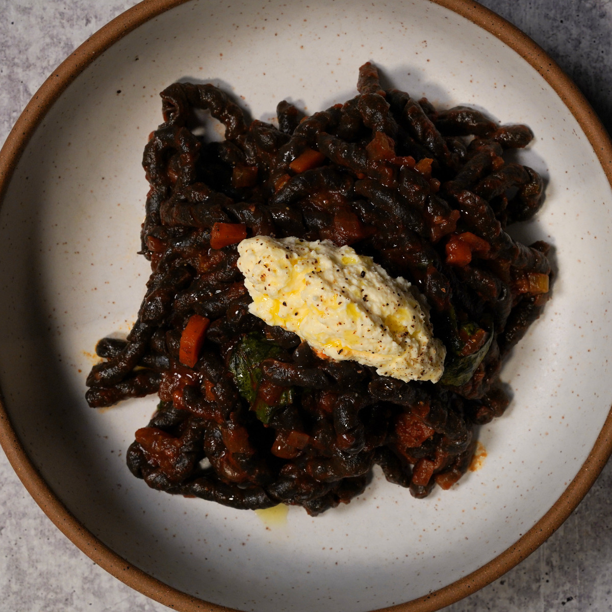 A plate of dark-colored pasta in a rich tomato-based sauce, topped with a dollop of creamy ricotta cheese and a drizzle of olive oil, served on a white ceramic dish with a brown rim.
