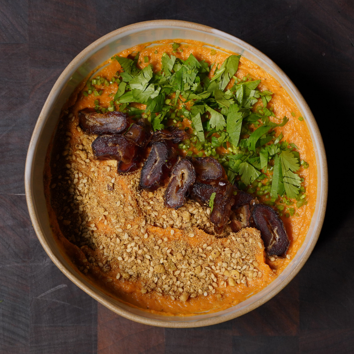 A bowl of orange-colored dip or spread, topped with chopped fresh herbs, crushed nuts, and sliced dates, arranged in neat sections. The bowl sits on a dark wooden surface.