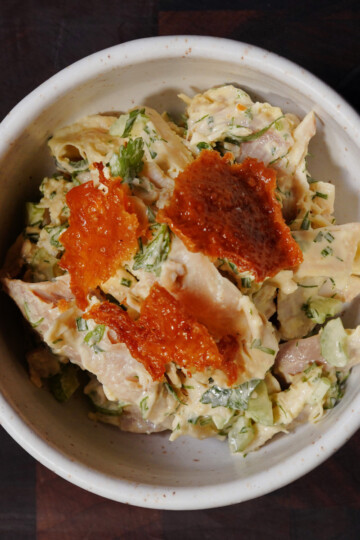 A bowl of chicken salad mixed with herbs and chopped celery, topped with three dollops of bright orange fish roe, served in a white ceramic bowl on a dark surface.