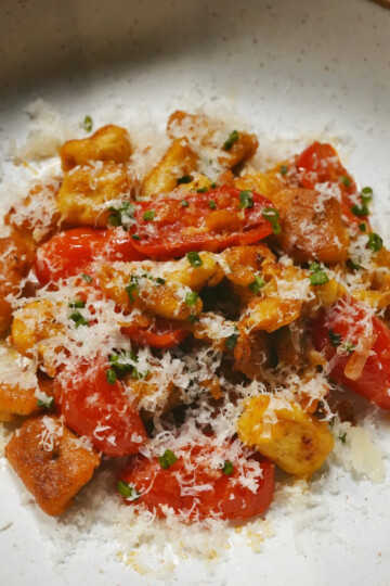 A bowl of gnocchi with roasted cherry tomatoes, topped with grated cheese and chopped herbs, served on a white ceramic plate with a brown rim.