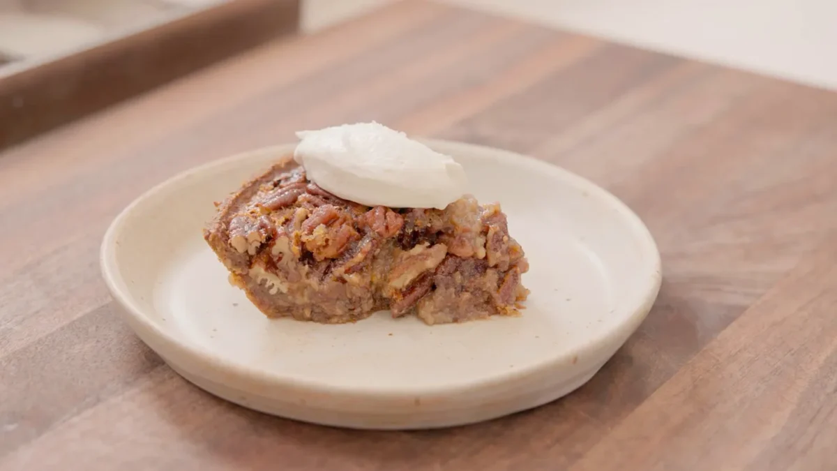 A slice of pecan pie sits on a round beige plate, topped with a dollop of whipped cream, on a wooden surface.