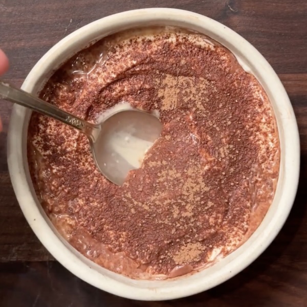 A bowl of chocolate oatmeal topped with cocoa powder, with a spoon scooping out a portion, revealing the creamy texture beneath. The bowl sits on a dark wooden surface.