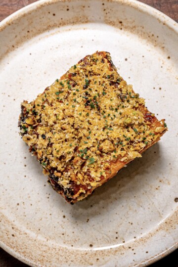A square slice of layered potato gratin topped with golden breadcrumbs and herbs, served on a rustic, speckled beige plate.