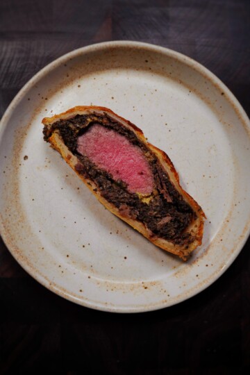 A single slice of beef Wellington, showing pink beef wrapped in mushroom duxelles and pastry, sits on a speckled white plate against a dark background.