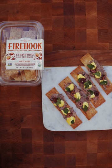 A container of Firehook Everything Like the Bagel organic crackers sits on a wooden surface next to a marble board with five crackers topped with spreads, sliced olives, herbs, and garnishes.