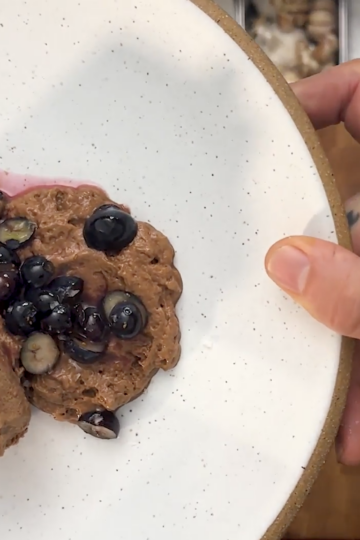 A hand holds a white plate with two slices of chocolate dessert topped with blueberries and sauce, viewed from above on a wooden surface.