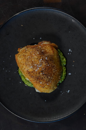 A piece of seasoned, roasted chicken with crispy skin sits on a dark plate atop a green sauce, likely pesto. The dish is garnished with flaky sea salt, and the background is a dark wood surface.