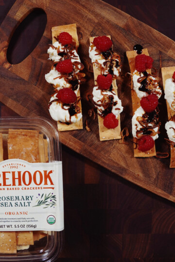 A wooden board holds five rosemary sea salt crackers topped with cream cheese, raspberries, and a drizzle of balsamic glaze. Next to it is a plastic container labeled &ldquo;Firehook Rosemary Sea Salt Crackers.&rdquo;.