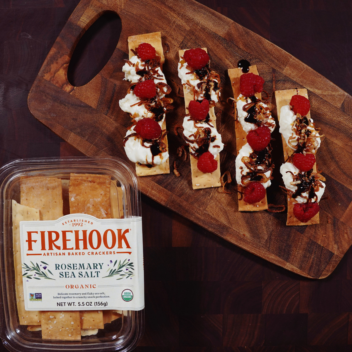 A wooden board holds five rosemary sea salt crackers topped with cream cheese, raspberries, and a drizzle of balsamic glaze. Next to it is a plastic container labeled 